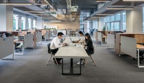 Modern office interior featuring a large open workspace with employees at various stations, highlighted by sleek wooden desks, partitions, and innovative overhead lighting fixtures.